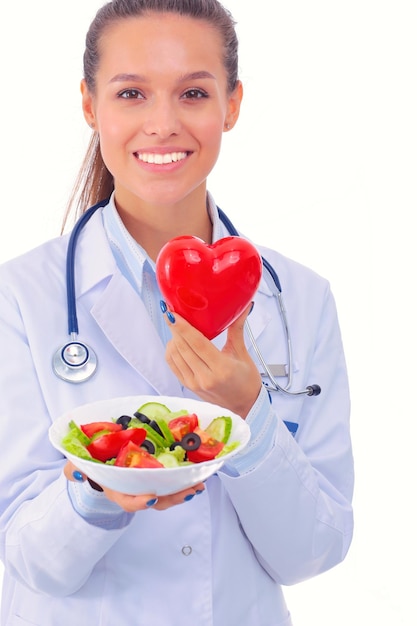 Portrait d'une belle femme médecin tenant une assiette avec des légumes frais et un coeur rouge