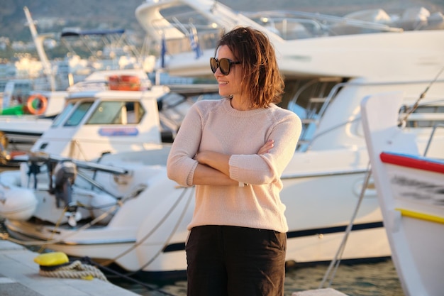 Portrait de belle femme mûre réussie, femme heureuse et confiante avec les bras croisés, baie de mer avec fond de coucher de soleil d'été de yachts