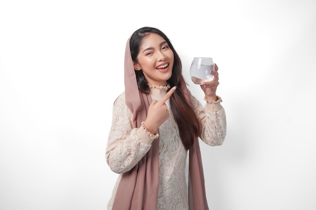 Portrait d'une belle femme musulmane asiatique en foulard, voile, hijab, souriante et montrant du doigt le verre d'eau qu'elle tient, concept du Ramadan