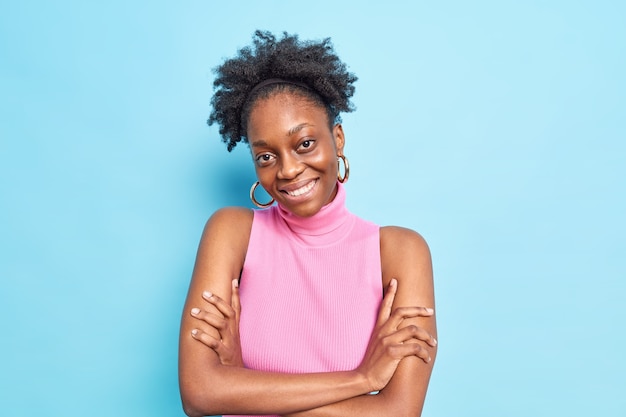 Portrait d'une belle femme à la peau foncée, les cheveux bouclés sourit, garde agréablement les bras croisés, incline la tête