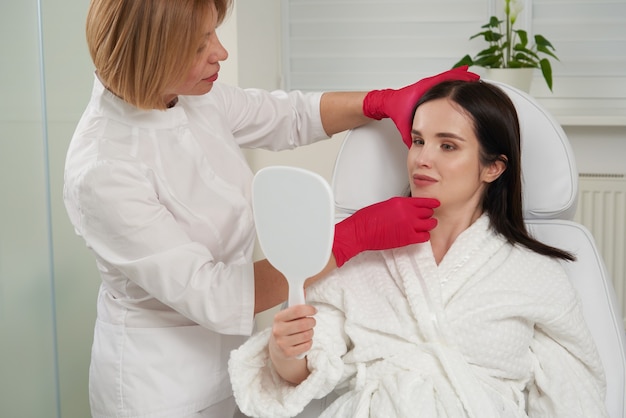 Portrait d'une belle femme en peignoir blanc assise sur le lit et vérifiant les résultats des soins de la peau