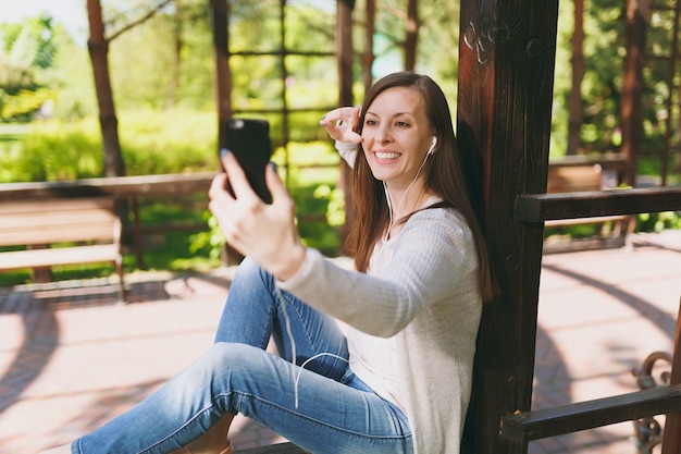 Portrait de belle femme portant des vêtements légers et décontractés. Femme souriante assise dans le parc de la ville dans la rue à l'extérieur sur la nature printanière, faisant une photo de selfie sur téléphone portable ou appel vidéo. Concept de mode de vie.