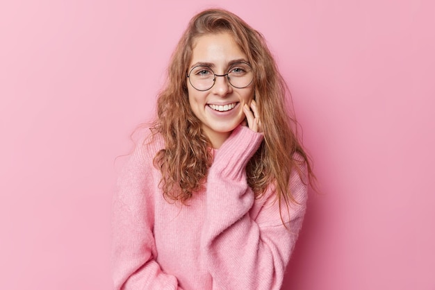 Portrait d'une belle femme positive avec de longs cheveux ondulés sourire à pleines dents garde la main sur les joues se sent heureuse d'avoir une conversation agréable porte un pull à manches longues lunettes rondes pose contre le mur rose