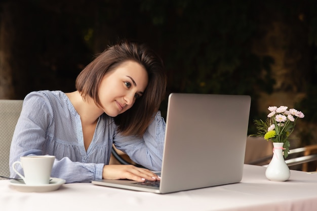 Portrait de la belle femme qui travaille à domicile, elle est assise avec une tasse de café à la table, travaillant sur un ordinateur portable à l'intérieur