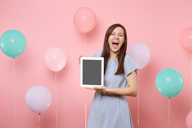 Portrait d'une belle femme ravie avec la bouche ouverte en robe bleue tenant un ordinateur tablette avec un écran vide vide sur fond rose avec des ballons à air colorés. Concept de fête d'anniversaire.