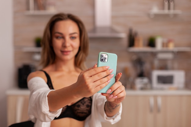 Portrait d'une belle femme regardant la caméra du téléphone tout en prenant un selfie dans la cuisine de la maison avec des rapports sexuels...