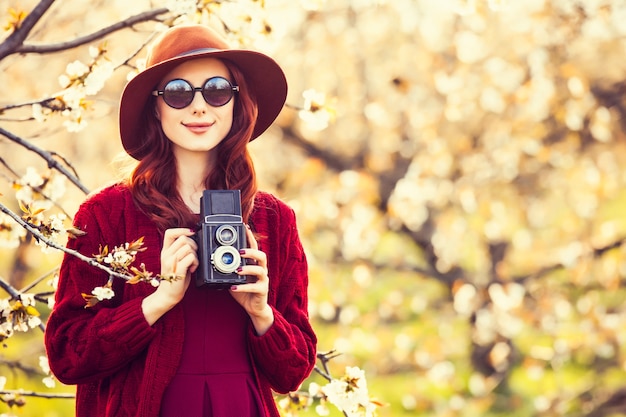 Portrait D'une Belle Femme Rousse En Pull Rouge Et Chapeau Avec Appareil Photo En Fleur De Jardin De Pommier Au Printemps Au Coucher Du Soleil.