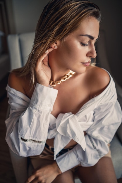 Portrait d'une belle femme séduisante aux cheveux blonds assise dans une pièce sur une chaise en chemise blanche et posant magnifiquement