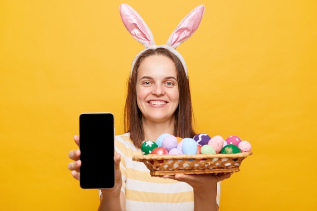 Portrait d'une belle femme souriante séduisante portant des oreilles de lapin tenant des oeufs de pâques et montrant un téléphone intelligent avec un espace de copie d'écran vide isolé sur fond jaune