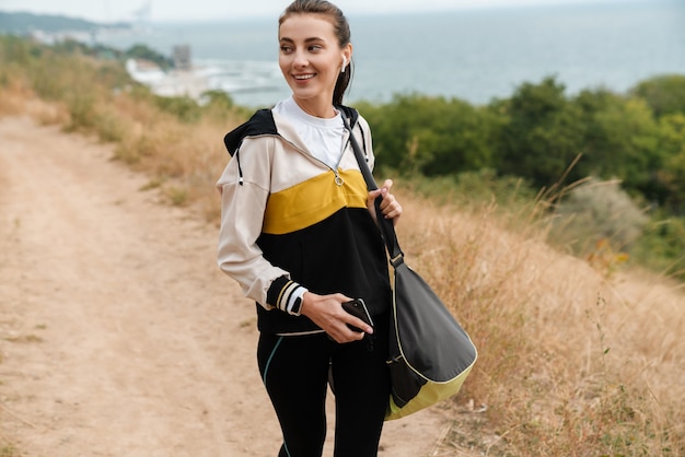 Portrait d'une belle femme souriante en tenue de sport utilisant des écouteurs et un téléphone portable tout en travaillant au bord de la mer