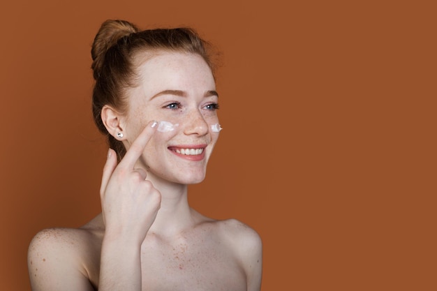 Portrait d'une belle femme souriante tout en appliquant de la crème pour le visage sur sa joue isolée sur le front...