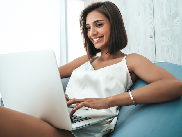 Portrait de belle femme souriante vêtue d'un pyjama blanc. Modèle insouciant assis sur une chaise souple et utilisant un ordinateur portable.