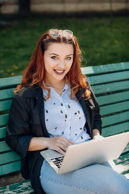 Portrait d'une belle femme tenant un ordinateur portable sur ses jambes alors qu'il était assis sur un banc à l'extérieur en souriant