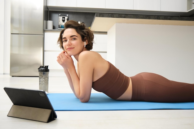 Portrait d'une belle femme en vêtements de sport de fitness se trouve sur le tapis de yoga en caoutchouc au sol utilise une tablette et un fil