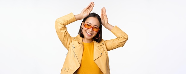 Portrait de belle fille asiatique à lunettes de soleil montrant le geste des mains des oreilles de lapin et souriant debout heureux sur fond blanc