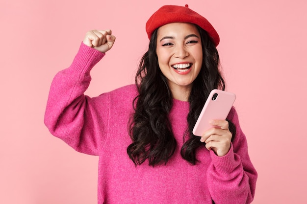 Portrait d'une belle fille asiatique optimiste portant un béret riant et tenant un smartphone isolé sur rose