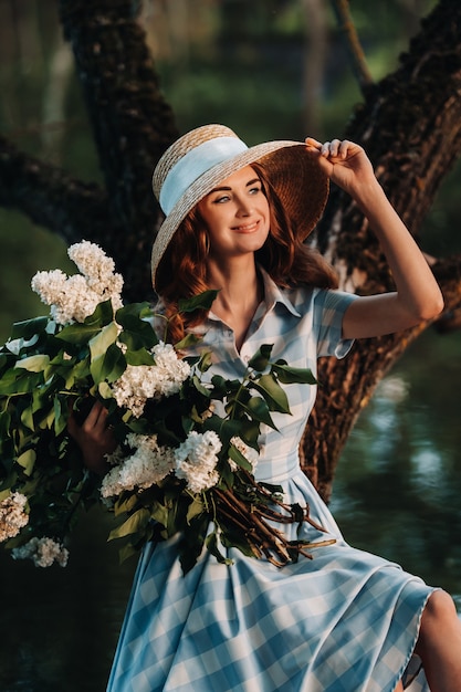 Portrait d'une belle fille aux cheveux longs, un chapeau de paille et une longue robe d'été avec des fleurs lilas dans le jardin