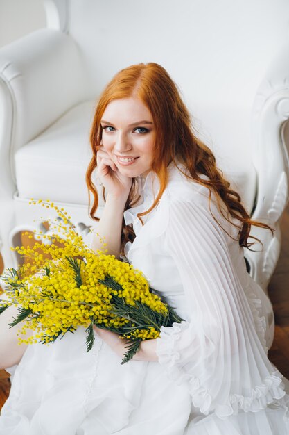 Portrait d'une belle fille aux cheveux roux avec un Mimosa dans une longue robe blanche