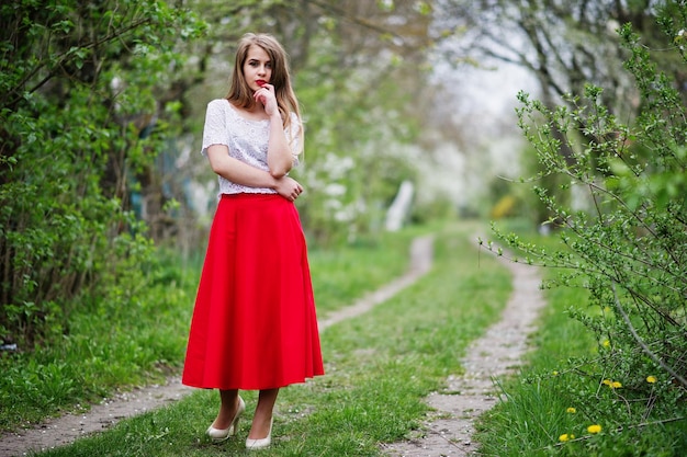 Portrait d'une belle fille aux lèvres rouges au jardin de fleurs de printemps portant une robe rouge et un chemisier blanc