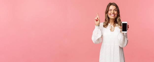 Portrait d'une belle fille blonde chanceuse et pleine d'espoir en robe blanche faisant un pari montrant l'affichage du téléphone portable doigt croisé bonne chance et souriante optimiste ayez foi qu'elle gagnera en compétition