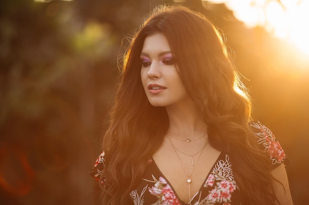 Portrait d'une belle fille brune aux cheveux épais ondulés contre un coucher de soleil de la ville Une fille en short jeans et un chemisier coloré noir avec un maquillage élégant et des bijoux