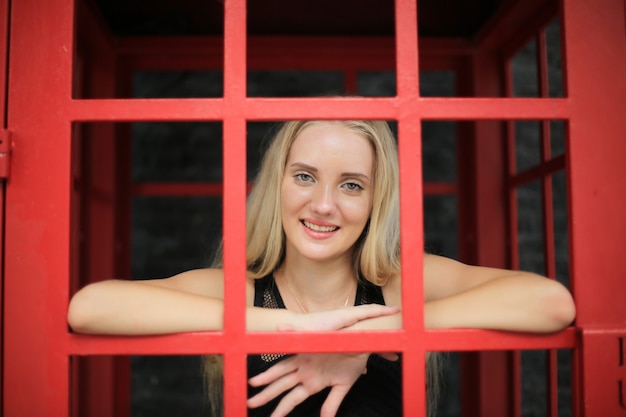 Portrait de belle fille de cheveux blonds sur la robe noire se tenant dans la cabine téléphonique rouge