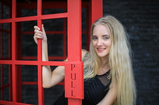 Portrait de belle fille de cheveux blonds sur la robe noire se tenant dans la cabine téléphonique rouge