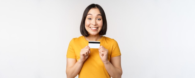 Portrait d'une belle fille coréenne tenant une carte de crédit recommandant un service bancaire debout en t-shirt jaune sur fond blanc