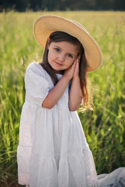 Portrait d'une belle fille dans un chapeau de paille debout dans un champ avec de l'herbe verte au coucher du soleil et en regardant la caméra