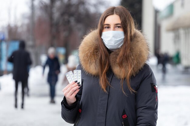 Portrait d'une belle fille dans un masque de protection médicale en tenant des pilules jaunes dans ses mains pour le rhume et la grippe Portrait de rue d'hiver d'une femme sous des chutes de neige