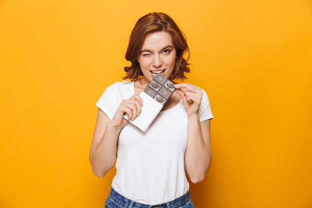 Portrait d'une belle fille heureuse debout isolée sur un mur jaune, tenant une barre de chocolat, mordant