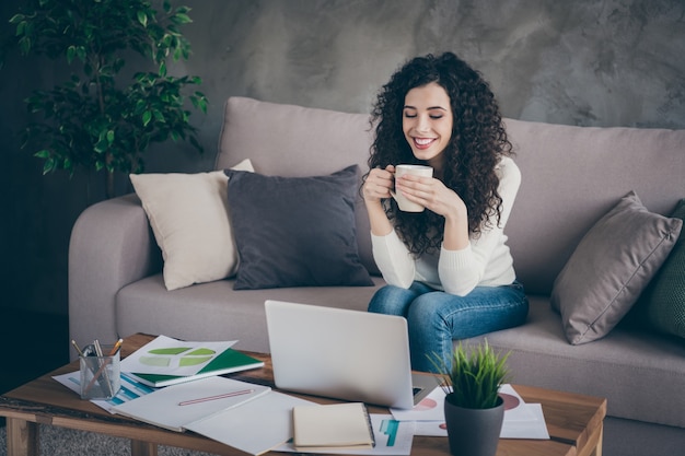 Portrait de belle fille joyeuse assise sur le canapé, boire du café, regarder la vidéo en ligne dans le salon intérieur de style industriel loft moderne à l'intérieur