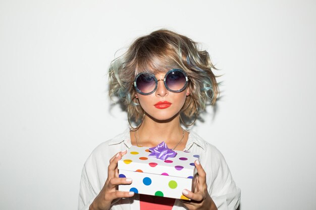 Portrait De Belle Fille En Lunettes De Soleil Et Boucles D'oreilles Regardant Rêveusement à Huis Clos Avec Boîte-cadeau Dans Les Mains Sur Fond Blanc Isolé