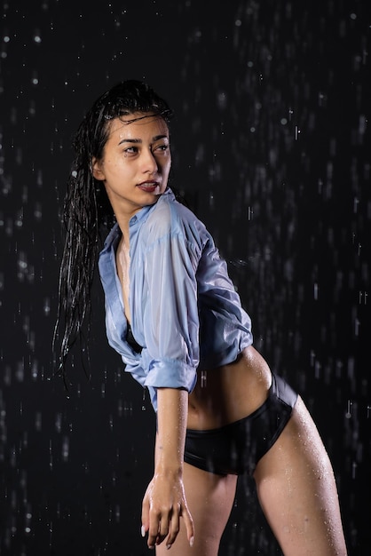 Portrait d'une belle fille en maillot de bain noir et chemise Debout sous la pluie en profitant des gouttes d'eau Éclaboussure d'eau sur fond noir Aqua studio