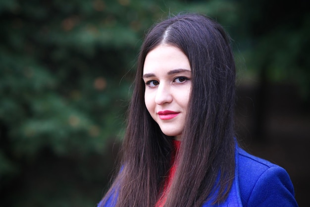 Portrait d'une belle fille de race blanche, jeune femme brune élégante et élégante aux cheveux longs et au manteau bleu regardant la caméra à l'extérieur, marchant dans le parc, la forêt