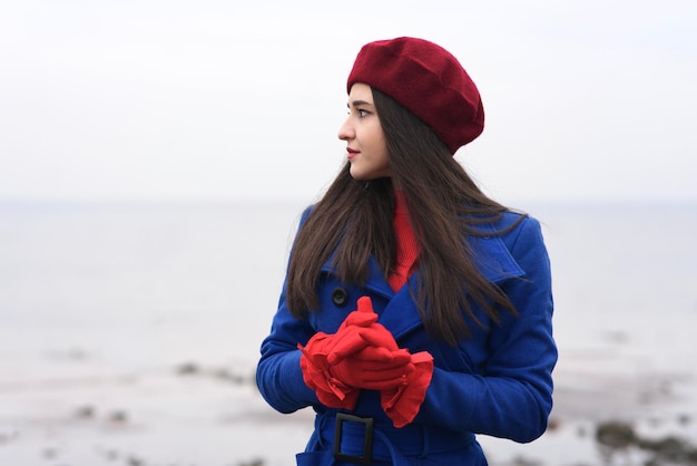 Portrait d'une belle fille réfléchie pensive, jeune femme brune élégante et élégante en béret rouge et gants et manteau bleu regardant à distance à l'extérieur, marchant sur la plage, l'océan.