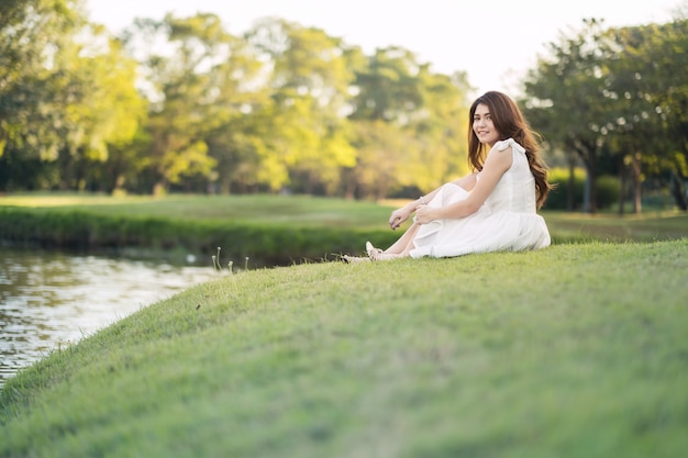 Portrait d'une belle fille en robe blanche, assise sur le gazon au bord de la rivière