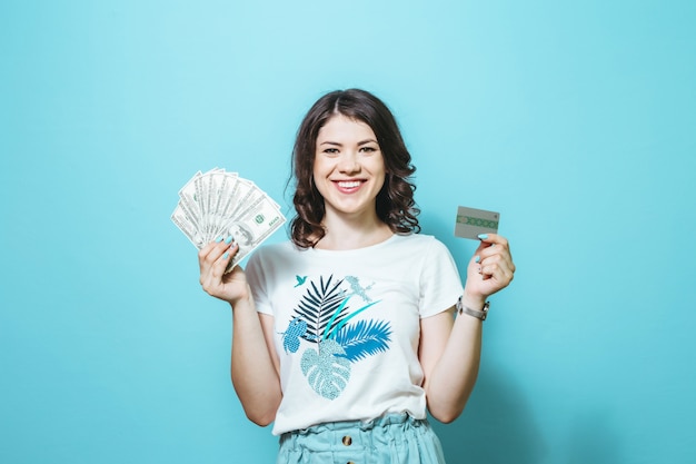 Portrait d'une belle fille satisfaite tenant des billets en argent et carte de crédit isolé sur fond bleu