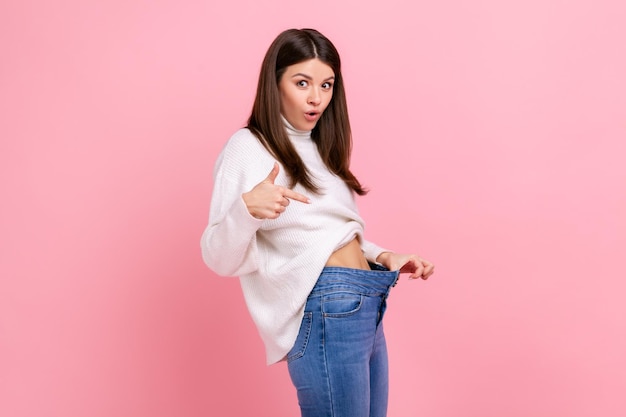 Portrait d'une belle fille surprise pointant vers une taille fine dans un gros pantalon perte de poids réussie