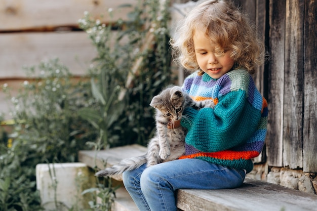 Portrait d'une belle et heureuse fille bouclée dans un pull tricoté jouant avec un chaton près d'un mur en bois