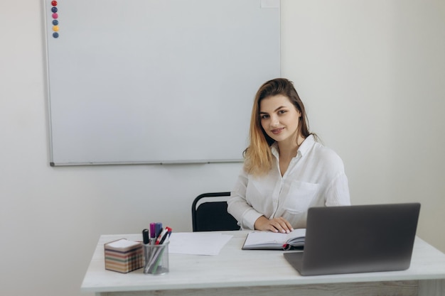 Portrait d'une belle jeune enseignante menant un apprentissage en ligne en classe pour ses élèves La fille est vêtue d'une chemise blanche Elle regarde la caméra