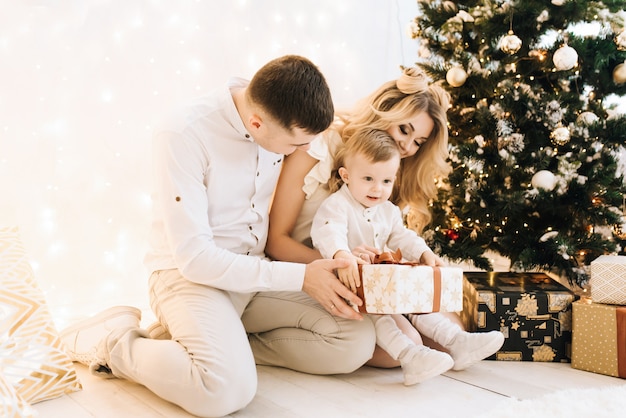 Portrait de la belle jeune famille sur l'arbre de Noël et fond de coton blanc. Des parents attrayants et un petit fils ouvrent les cadeaux du nouvel an