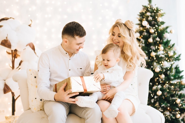 Portrait de la belle jeune famille sur l'arbre de Noël et fond de coton blanc. Des parents attrayants et un petit fils ouvrent les cadeaux du nouvel an