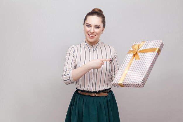 Portrait d'une belle jeune femme adulte satisfaite dans un style décontracté debout et pointant le doigt pour présenter avec un arc jaune et un sourire à pleines dents, regardant la caméra. Intérieur, isolé, tourné en studio, fond gris