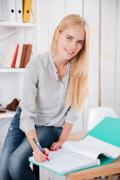 Portrait d'une belle jeune femme d'affaires assise sur un bureau et prenant des notes dans des documents