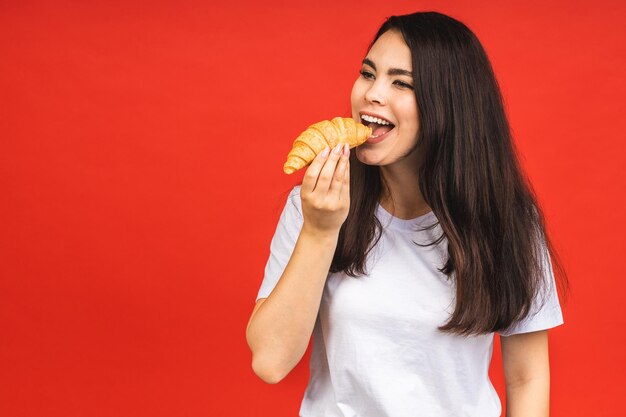 Photo portrait d'une belle jeune femme affamée mangeant un croissant portrait isolé d'une femme avec restauration rapide sur fond rouge concept de petit-déjeuner diététique