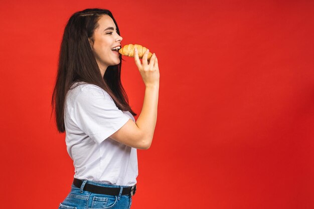 Portrait d'une belle jeune femme affamée mangeant un croissant Portrait isolé d'une femme avec restauration rapide sur fond rouge Concept de petit-déjeuner diététique