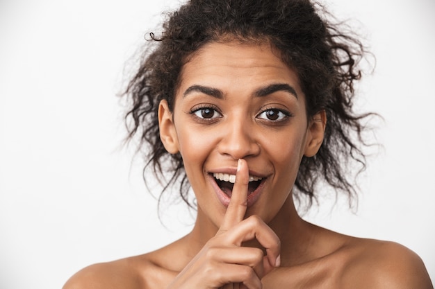 Portrait d'une belle jeune femme africaine heureuse posant isolé sur un mur blanc montrant le geste de silence.