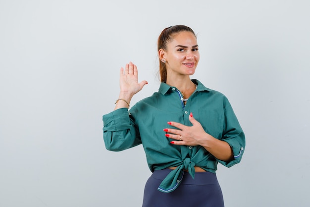 Portrait d'une belle jeune femme agitant la main pour saluer tout en gardant la main sur la poitrine en chemise verte et en regardant joyeuse vue de face