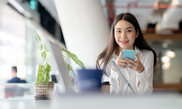 Portrait de la belle jeune femme asiatique à l'aide de smartphone au bureau, souriant et regardant la caméra.